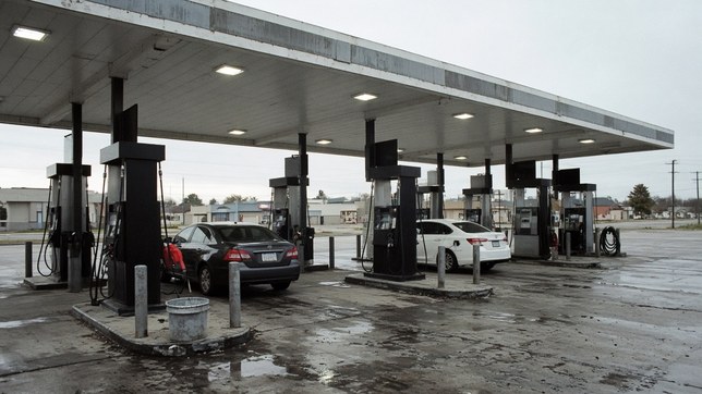 An American fuel forecourt under an overcast sky, two cars filling and the remaining pumps standing empty on wet concrete