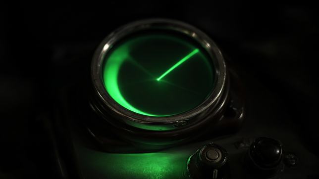 A circular radar scope glowing green in a darkened room, the sweep line partway round its arc with the trail fading behind it and the screen otherwise empty.