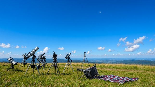 A row of telescopes and cameras on a sunlit grassy ridge aimed at one bright point high in a clear blue sky, a tipped camp chair and blanket beside them