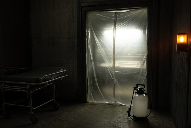 The sealed doorway of an isolation ward, heavy translucent plastic sheeting drawn taut across the frame and lit hard from the room beyond, an empty gurney turned aside in the dark and a chlorine sprayer standing on the concrete floor beneath an amber warning lamp.