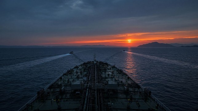 The bow of an oil tanker running through a strait at dusk, two small boats crossing ahead of it under an orange sunset behind dark headlands