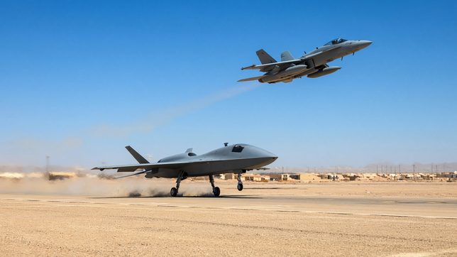 A gray uncrewed combat jet rolling down a desert runway kicking up dust while a crewed fighter banks low overhead against a clear blue sky