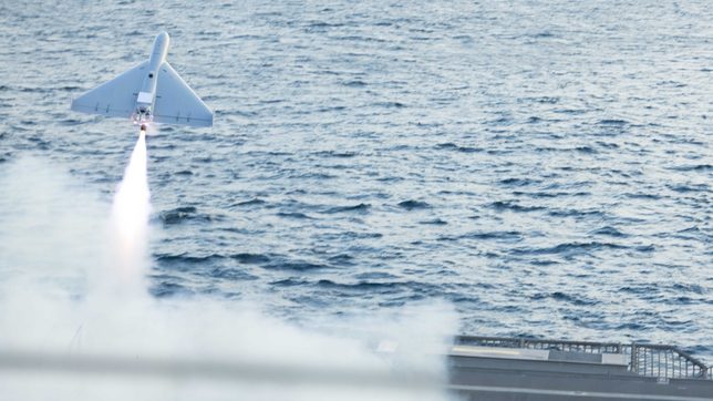 A one-way attack drone climbing away from a warship deck over open sea, its rocket booster burning a bright trail through the smoke