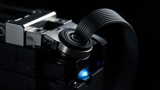 A robotic servo joint mid-articulation in machined aluminium and black polymer, ribbon cable looping away, a cold blue light deep in the housing.