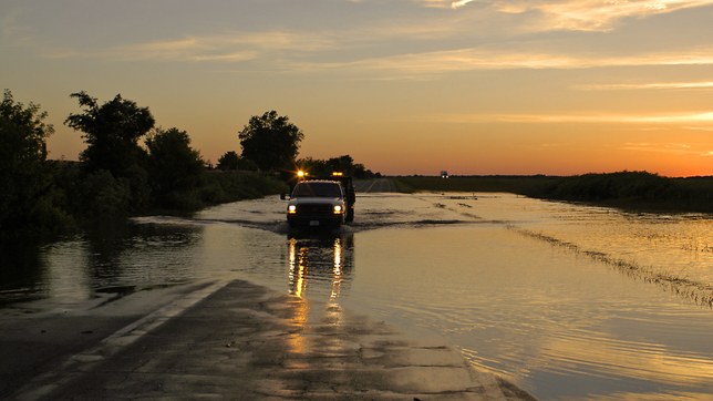 A rural highway submerged at sunset, a county road truck driving through the standing water with its beacons lit, floodwater lying across the fields on both sides